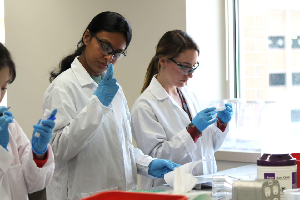 Students in white coats, goggles, and gloves work on an experiment in lab