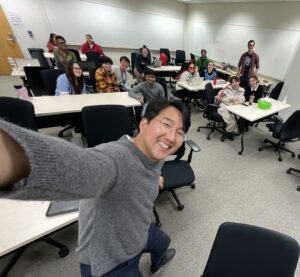 Dr. Hong taking a selfie with Hong research members sitting at tables behind him with some in their Halloween costumes