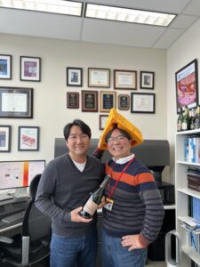Dr Nam with cheese hat on and and Dr Hong with bottle of champaign in office