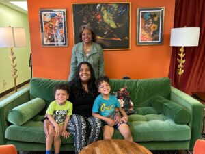Prof. Eva Vivian in front of a vibrant orange wall, standing over a green couch that holds Tracy Russell and her grandkids