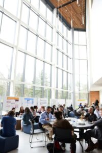 A convention with various people sitting down at tables eating and various studies printed onto tri-folds, lined along the windows for the MIKIW Medicinal Chemistry Conference