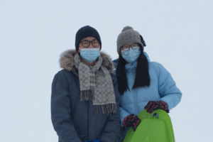 Suhjung Janet Lee and Zhen Zheng in coats with hats and gloves on while holding sleds