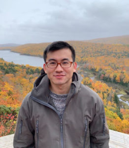Yuhui Li outdoors with colorful fall trees behind