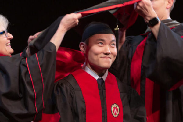 A pharmacy student stands smiling as two professors put on his hood at graduation