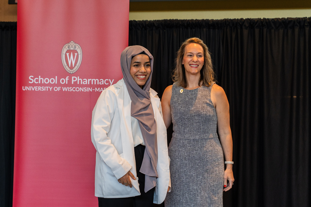 A student poses with Kate Rotzenberg in front of a red School of Pharmacy banner