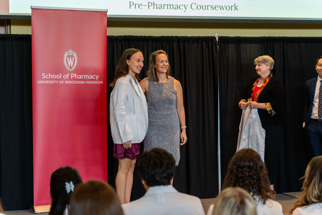 A student poses with Kate Rotzenberg in front of a red School of Pharmacy banner while Karen Kopacek holds a coat awaiting the next student