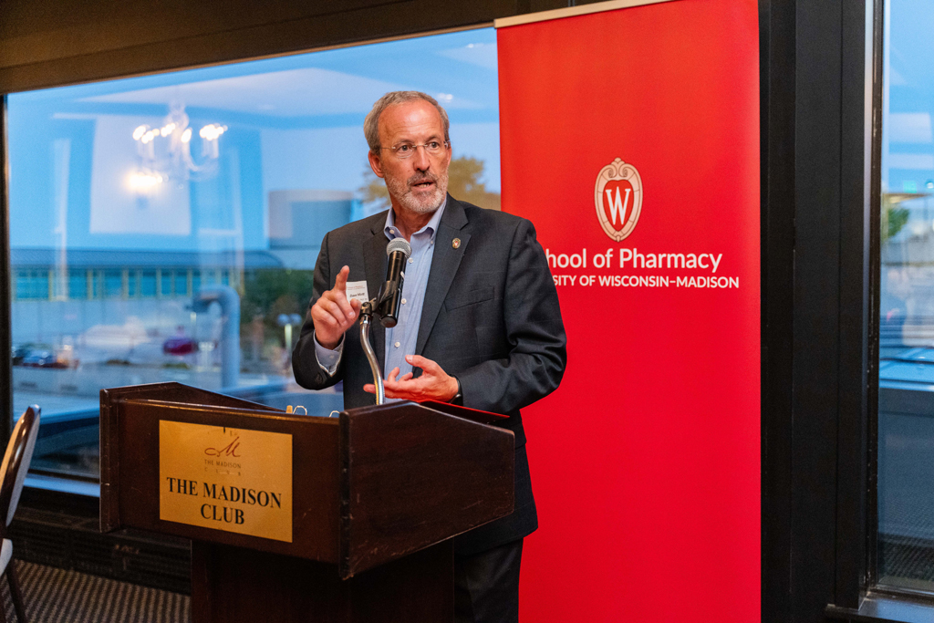 Professor Dave Mott speaks at the podium in front of a red School of Pharmacy background.