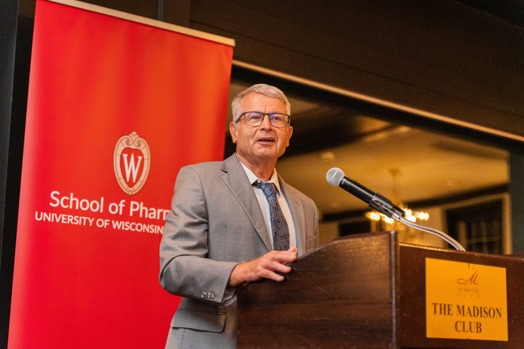 Evgenyi Shalaev speaks at a microphone in front of a red School of Pharmacy banner.