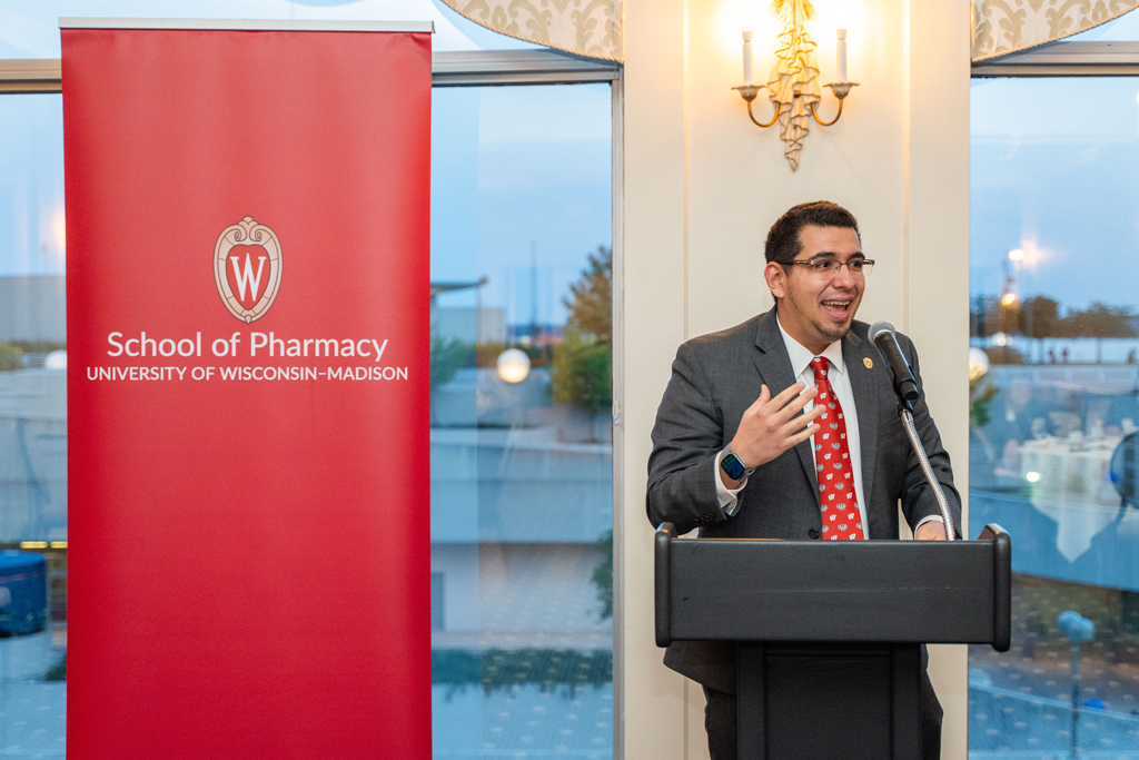 Ed Portillo speaks into a microphone podium in front of a red School of Pharmacy banner