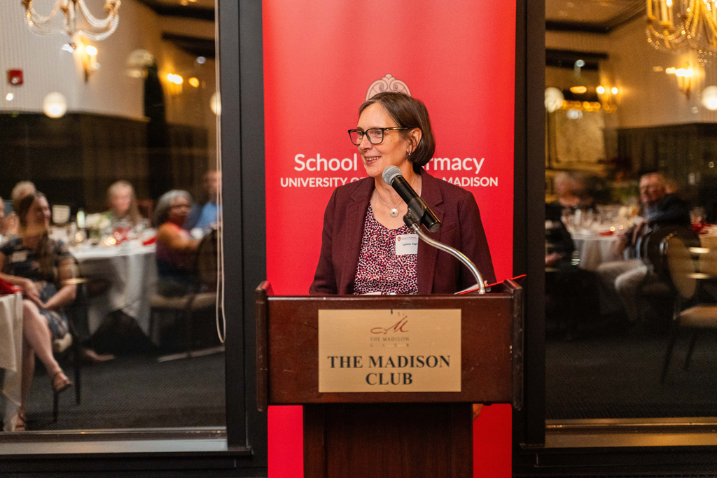 Lynne Taylor speaks at a microphone in front of a red School of Pharmacy banner.