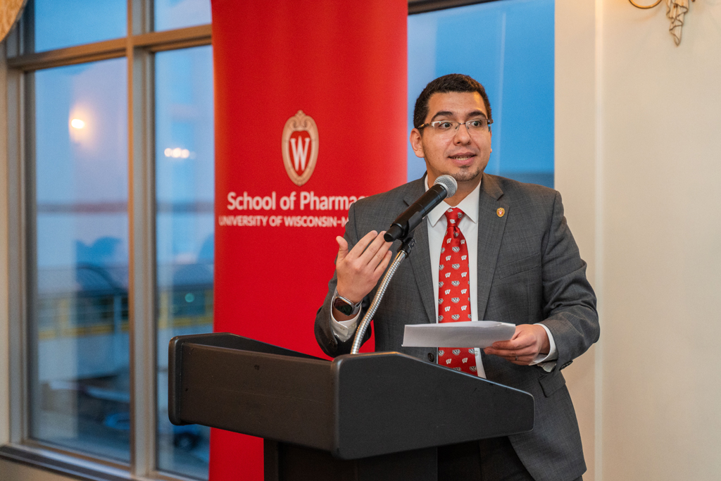 Ed Portillo speaks into a microphone podium in front of a red School of Pharmacy banner