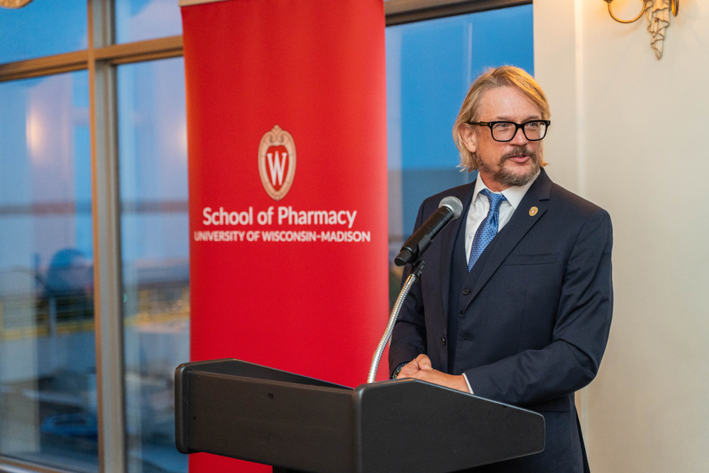 John Hill speaks into a microphone at a podium in front of a School of Pharmacy banner