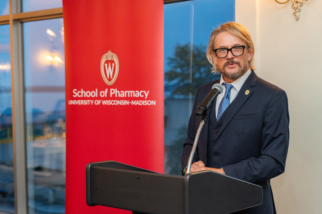 John Hill speaks at a podium in front of a red School of Pharmacy banner