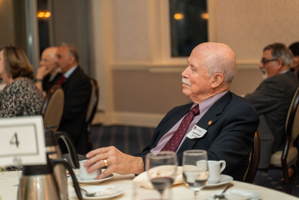 A man in a dark suit with a red tie and a nametag is listening to a speaker.