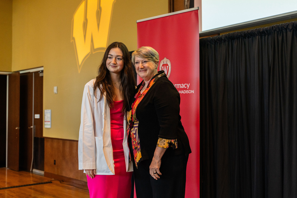 A student in a white coat poses with Karen Kopacek in front of a red School of Pharmacy banner