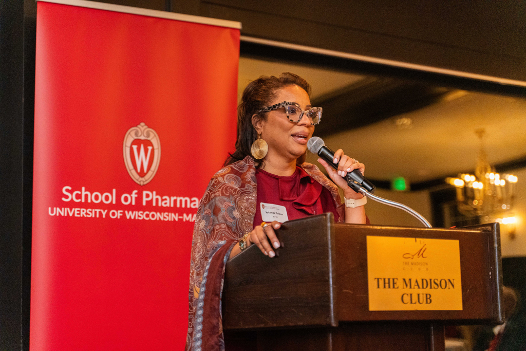 Yolanda Tolson speaks at a microphone in front of a red School of Pharmacy banner.