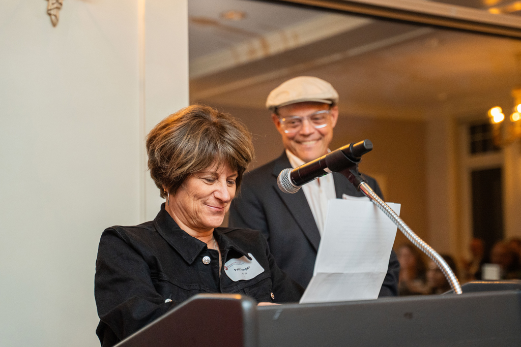 Patti Langer speaks at a microphone while Jeff Langer looks on and smiles