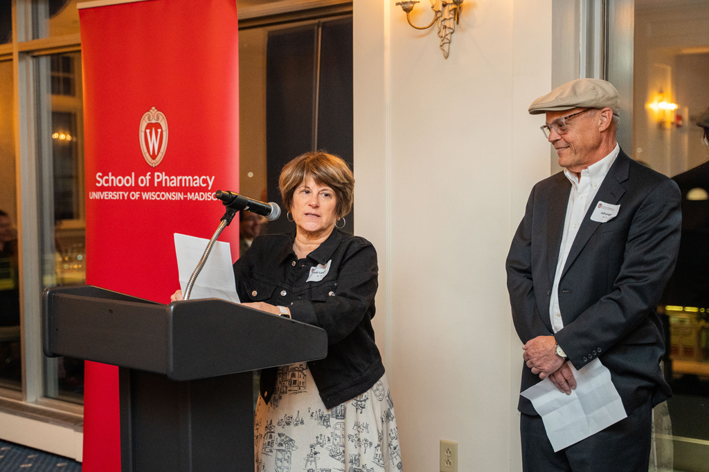 Patti Langer speaks into a podium microphone while Jeff Langer looks on