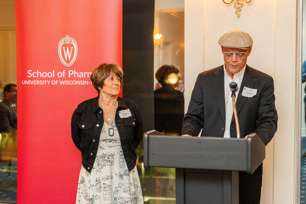 Jeff Langer speaks at a microphone in front of a red School of Pharmacy banner while Patti Langer looks on