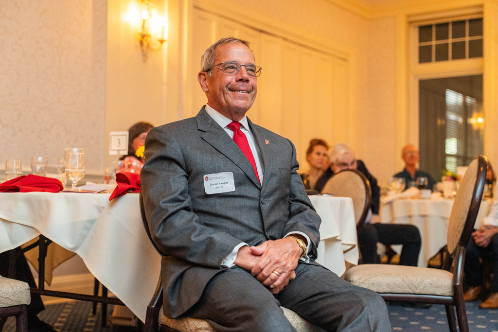 A man in a gray suit, Daniel Langer, smiles while seated at his table
