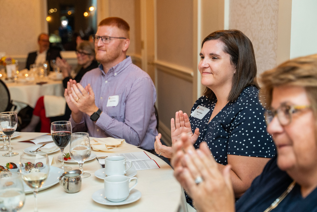 A man and a woman, Andrea Kuban, are seated at a dinner table with silverware, wine glasses, and coffee cups, smiling and clapping along with the other attendees at the event.