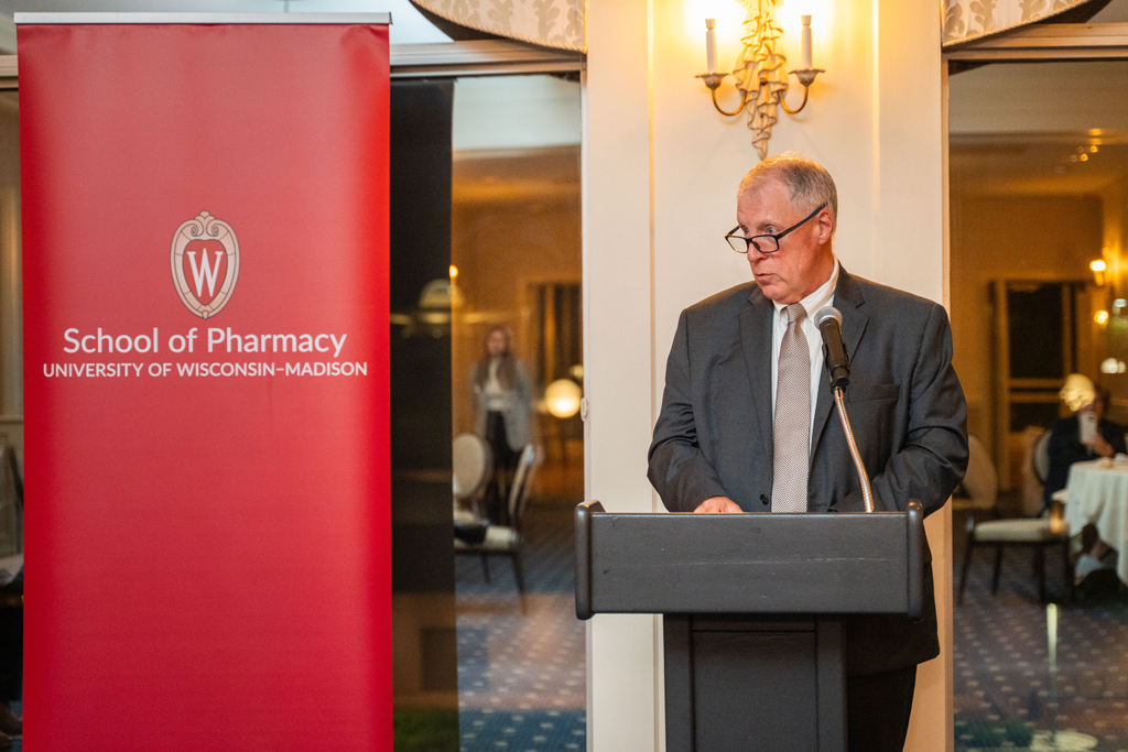 An older man in a dark gray suit and tie is speaking seriously at a black podium in an event room with decorative wall sconces. A large red banner for the School of Pharmacy is behind him.