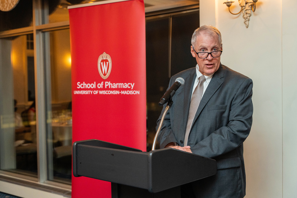 An older man in a dark gray suit and tie with glasses is speaking at a black podium with his mouth slightly open, appearing to deliver a speech. A large red banner for the School of Pharmacy is behind him.