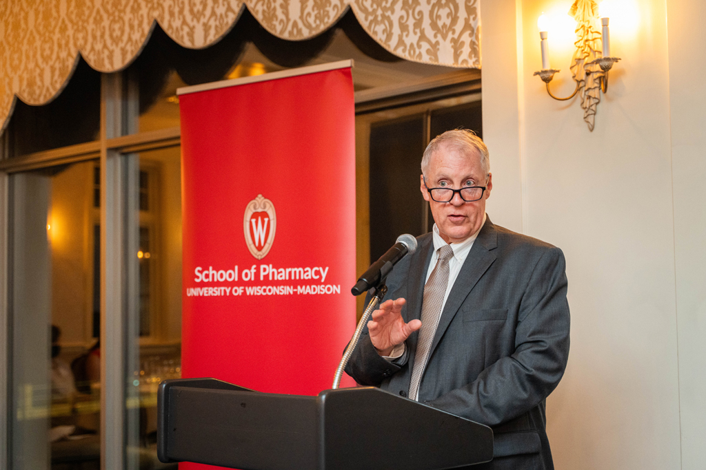 Eugene McNally speaks at a microphone in front of a red School of Pharmacy banner