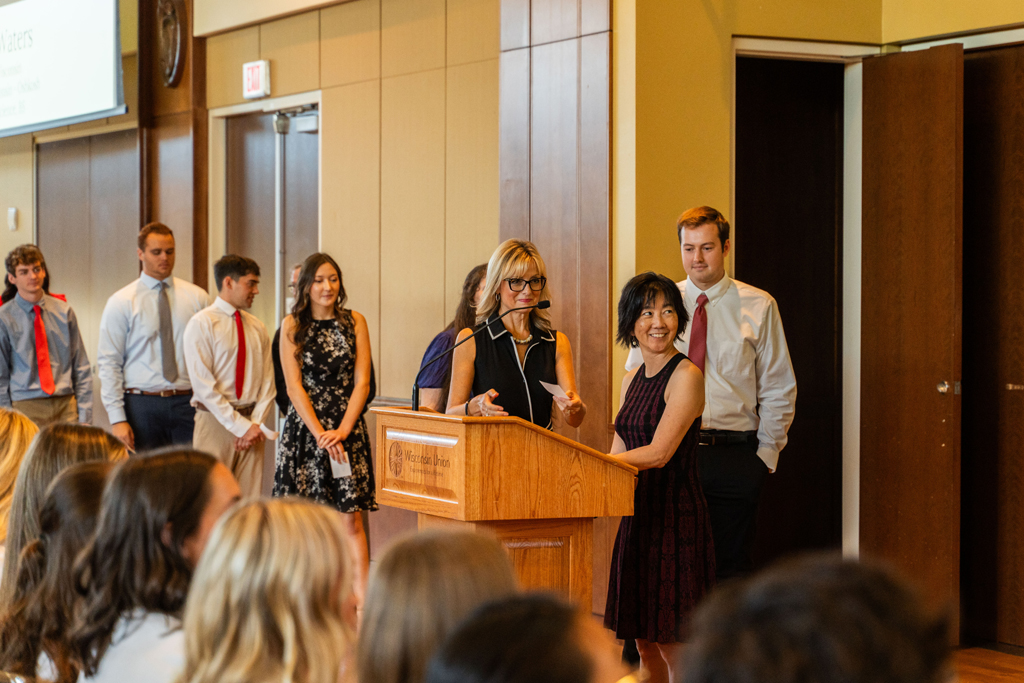 Faculty stand at a podium, students lined up behind them waiting for their white coats.