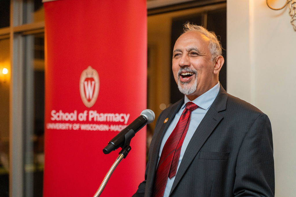 A man in a dark gray suit and a red tie, with a gray goatee, is standing at a microphone and smiling broadly, appearing to be laughing or delivering a humorous line during a speech.