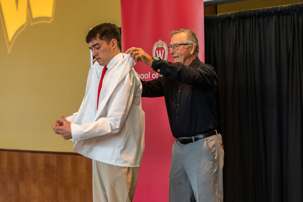 An older man in a black shirt places a white coat on a male student