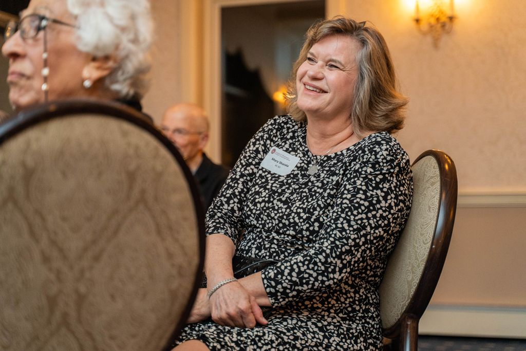 A woman, whose nametag reads Mary Schultz, is seated at a table, smiling and looking up and to the right of the camera.