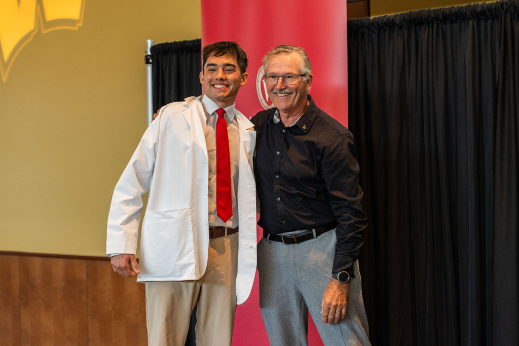 A student in a white coat and an older man smile and pose in front of a red School of Pharmacy banner