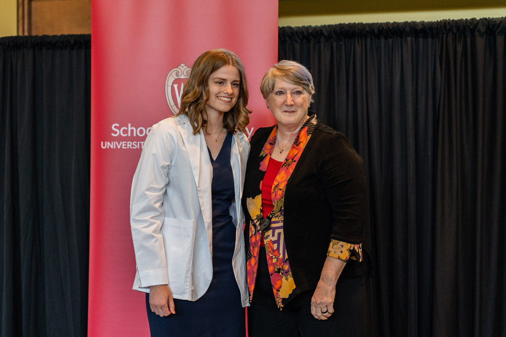 A student in a white coat poses with Karen Kopacek in front of a red School of Pharmacy banner