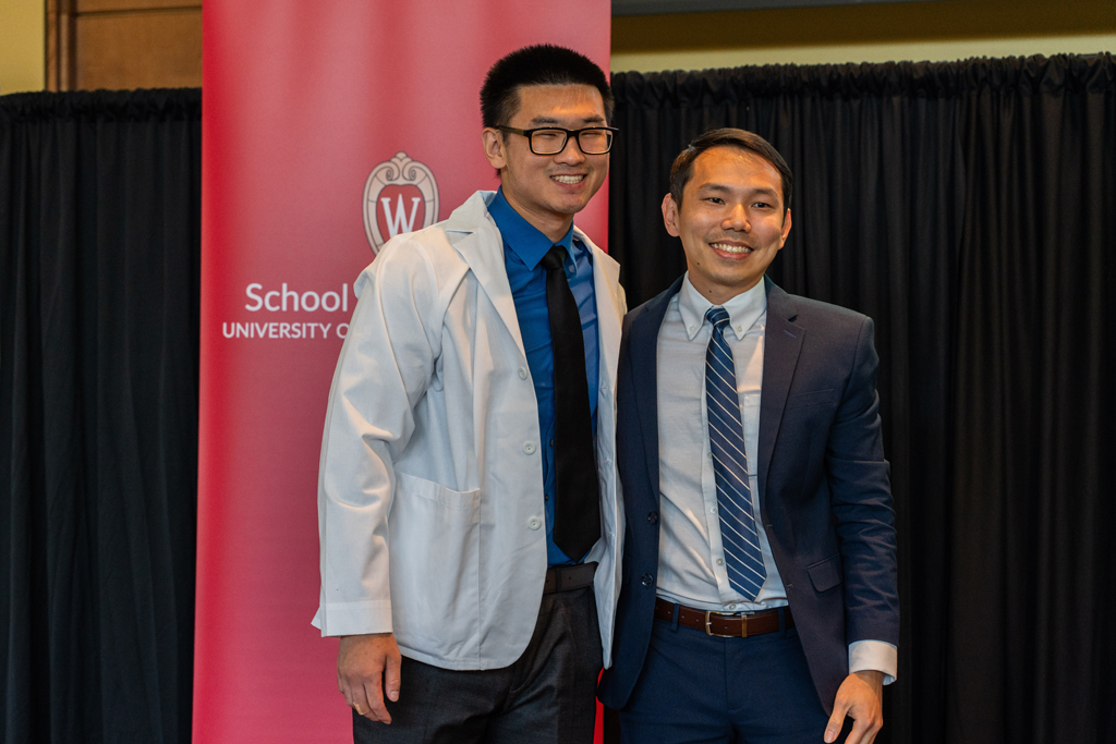 A student in a white coat poses with Sean Lim in front of a red School of Pharmacy banner