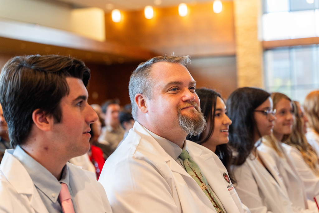 Seated students in white coats smile and listen to a speaker