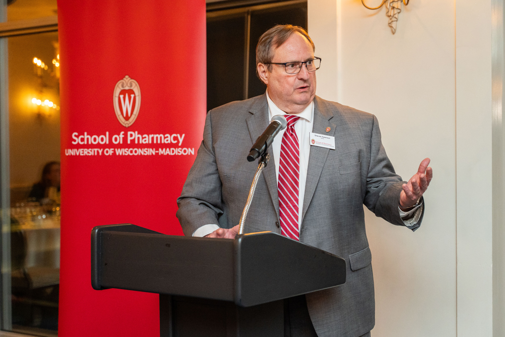 A man in a gray suit and a red-and-white striped tie, Steve Swanson, is speaking at a black podium and gesturing with his right hand. A large red banner for the School of Pharmacy is visible to his left.