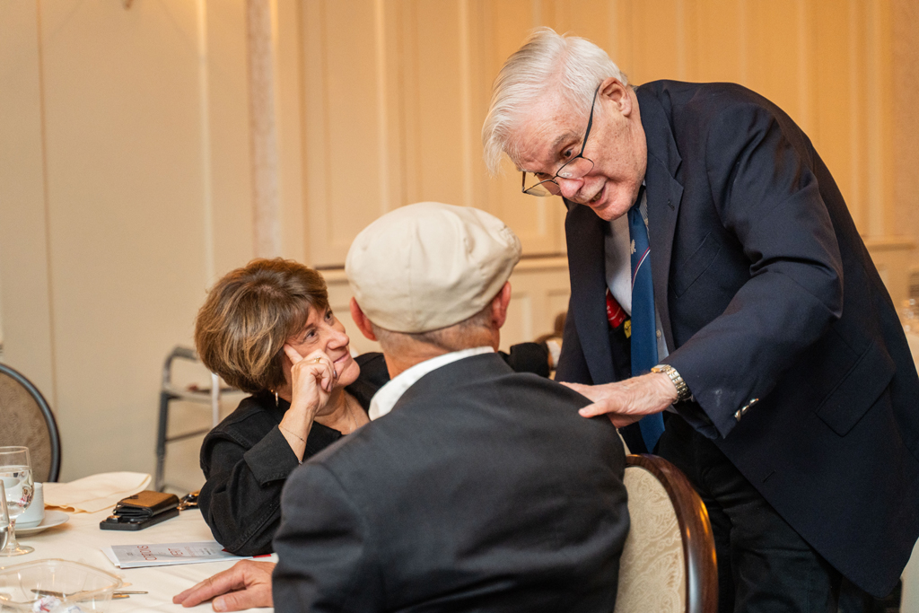 A woman and an older man in a navy suit are seated at a table, engaged in a close conversation with a third man whose back is to the camera.