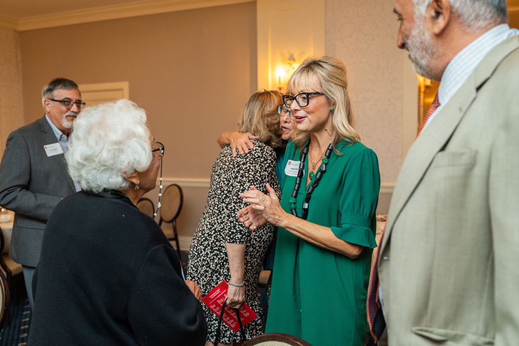 Several people are chatting in a formal event space. In the foreground, a woman in a bright green dress and glasses is speaking and gesturing with her hands.