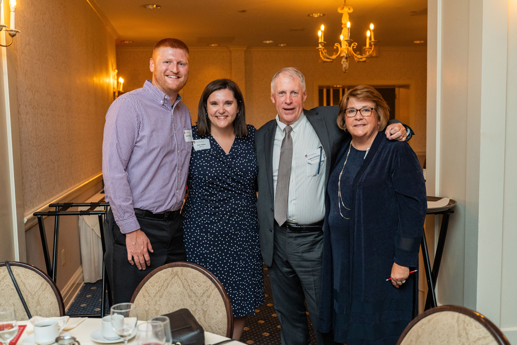 our people are standing together and smiling for a formal photo. From left to right: a man, a woman in a navy polka-dot dress whose nametag reads Andrea Kuban, an older man in a suit, and an older woman in a navy dress.