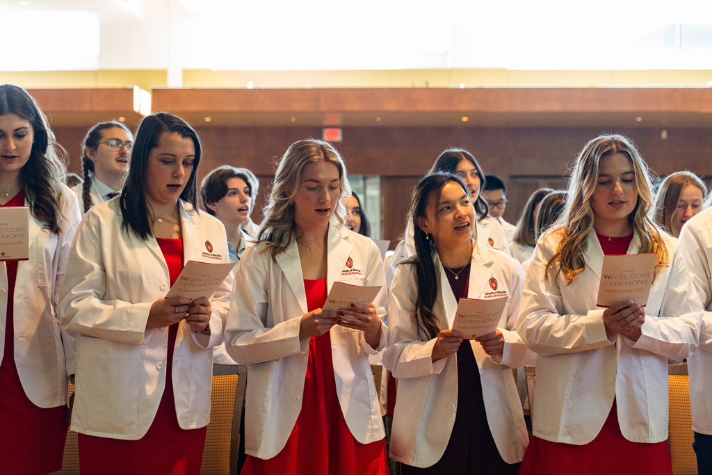 Students in white coats read the Oath of a Pharmacist off the backs of their ceremony programs