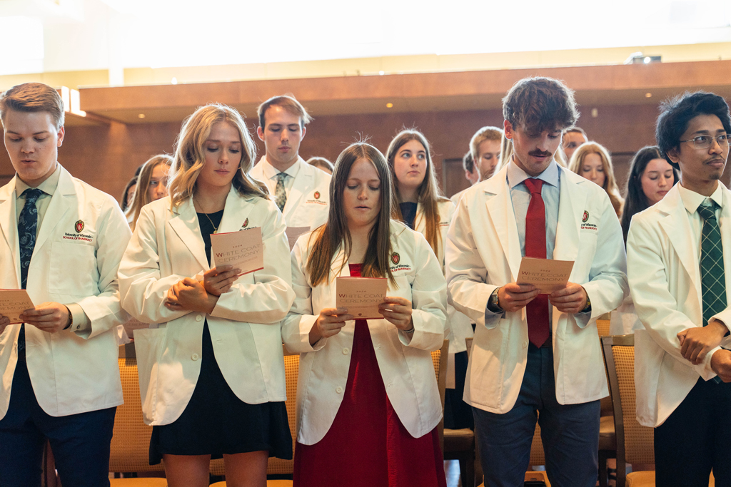 Students in white coats read the Oath of a Pharmacist off the backs of their ceremony programs