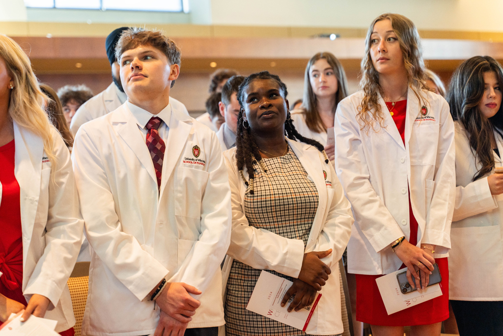 Standing students in white coats listen in to a speaker