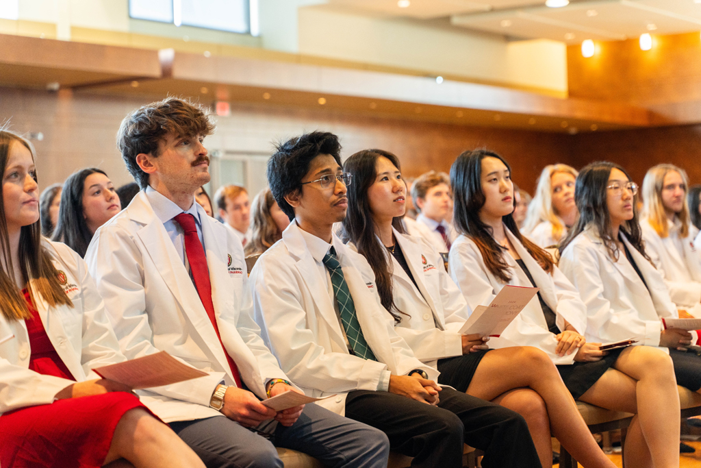 Seated students in white coats listen in to a speaker