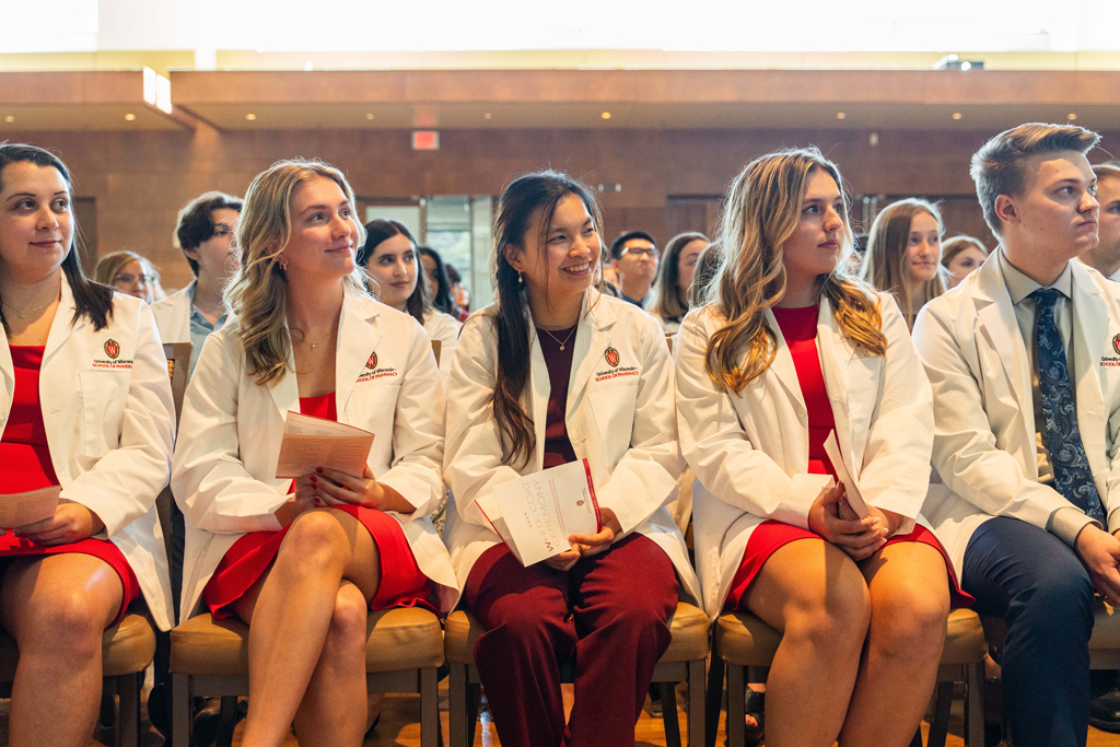 Seated students in white coats smile and listen in to a speaker