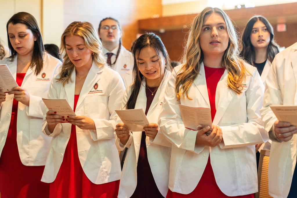 Students in white coats read the Oath of a Pharmacist off of screens in front of the room