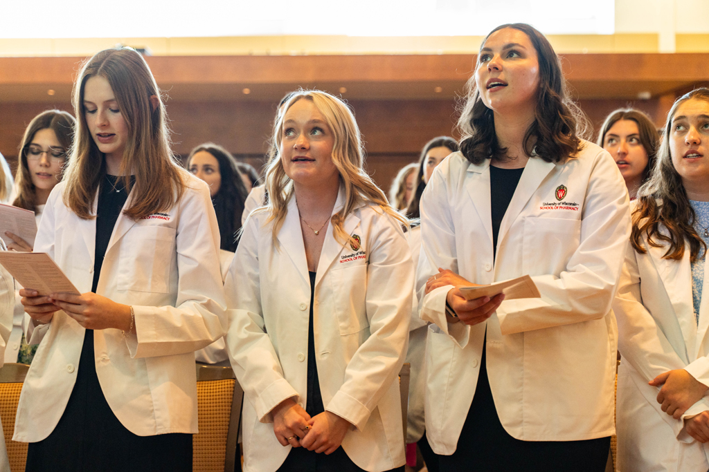 Students in white coats read the Oath of a Pharmacist off of screens in front of the room