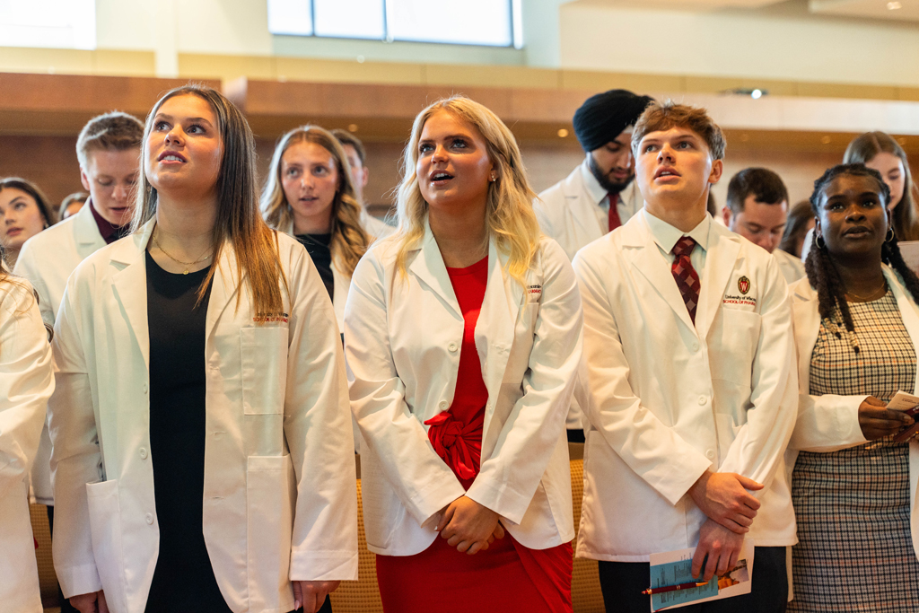 Students in white coats read the Oath of a Pharmacist off of screens in front of the room