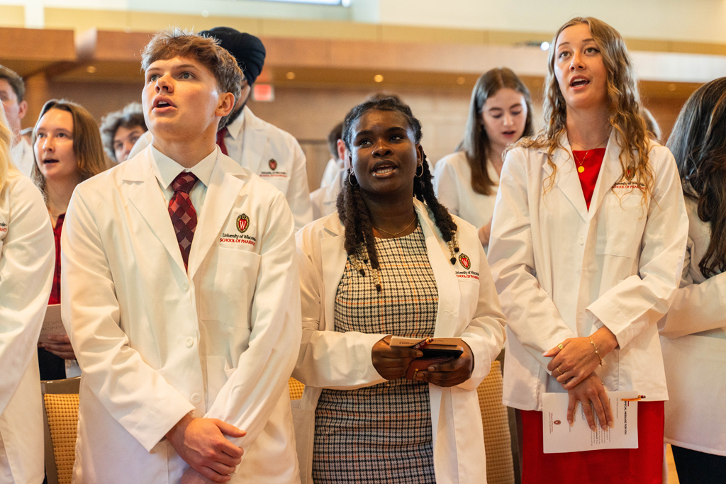 Students in white coats read the Oath of a Pharmacist off of screens in front of the room