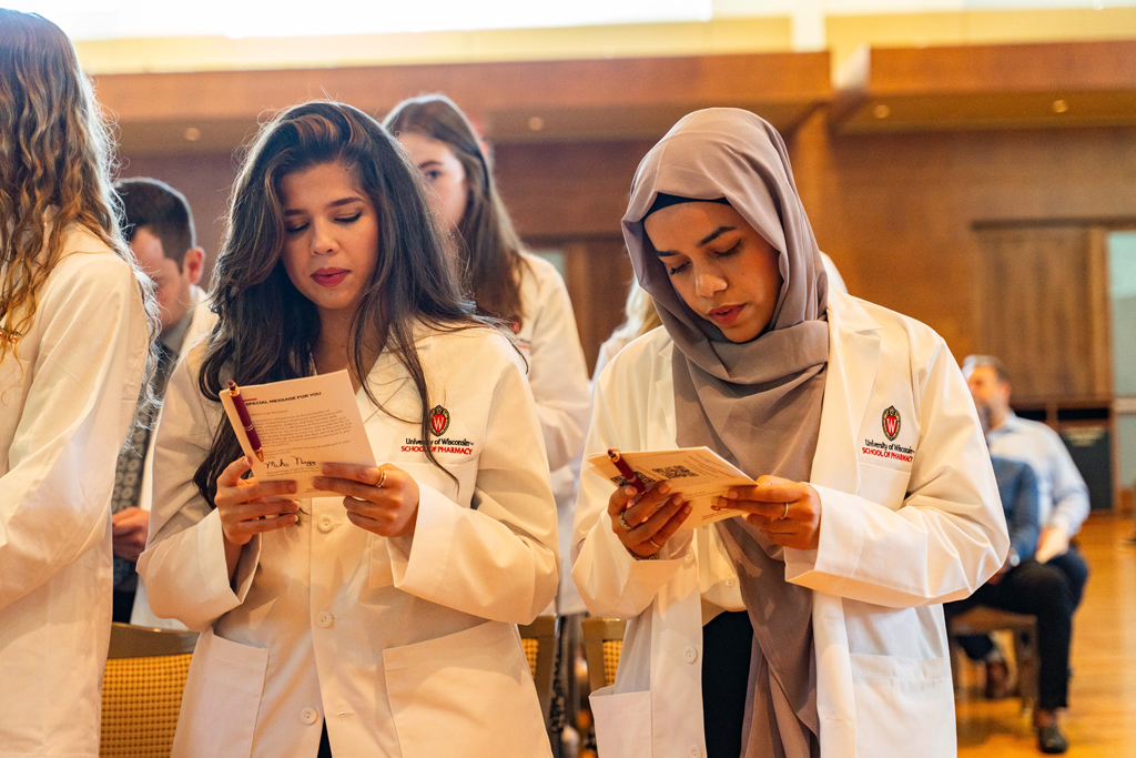 Female students in white coats read the Oath of a Pharmacist off the back of their White Coat Ceremony program